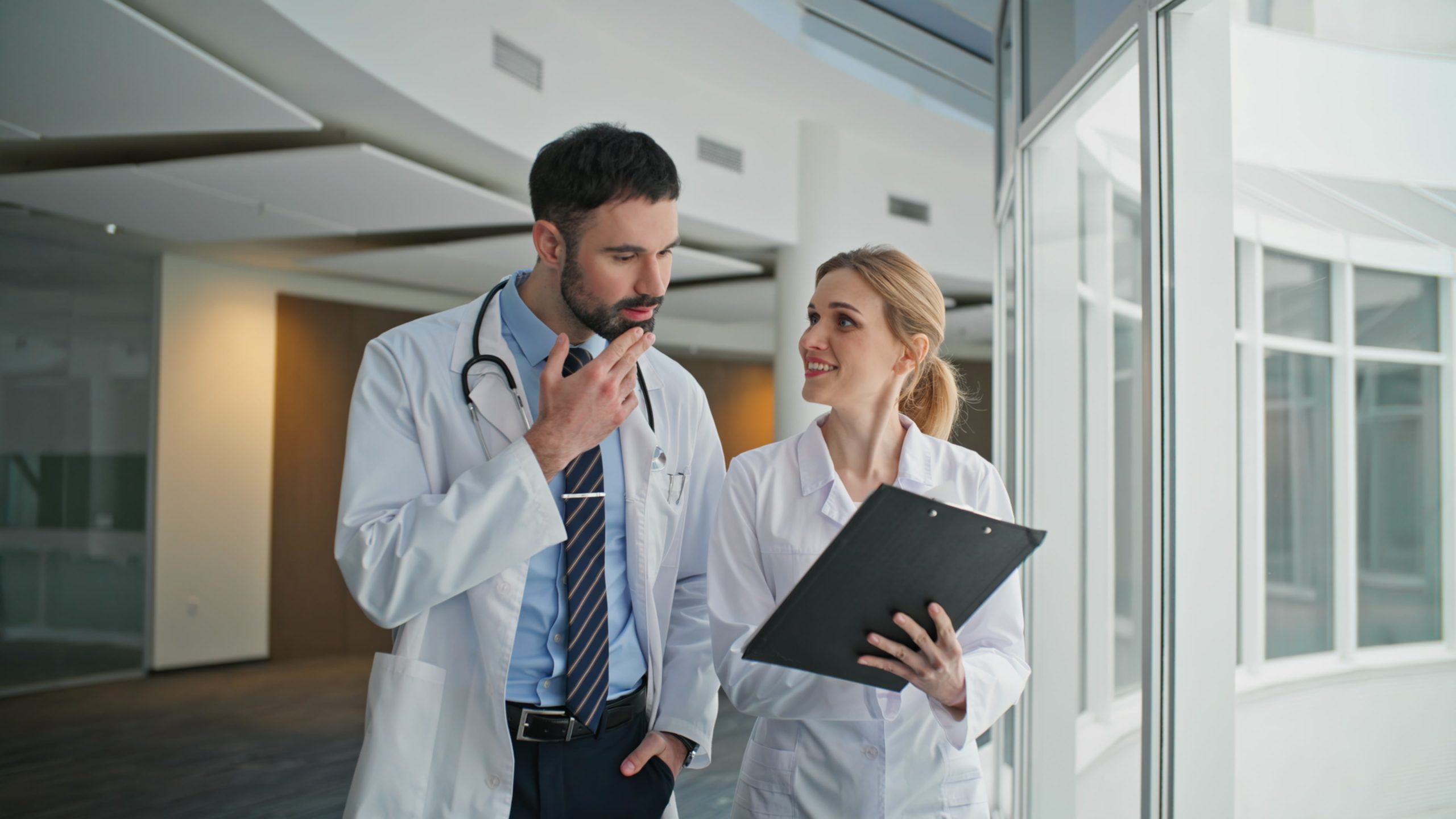Medical staff discussing clipboard chart in clinic. Smiling doctor talking nurse walking in hospital corridor. Healthcare professionals showing determination. Medical experts checking test results