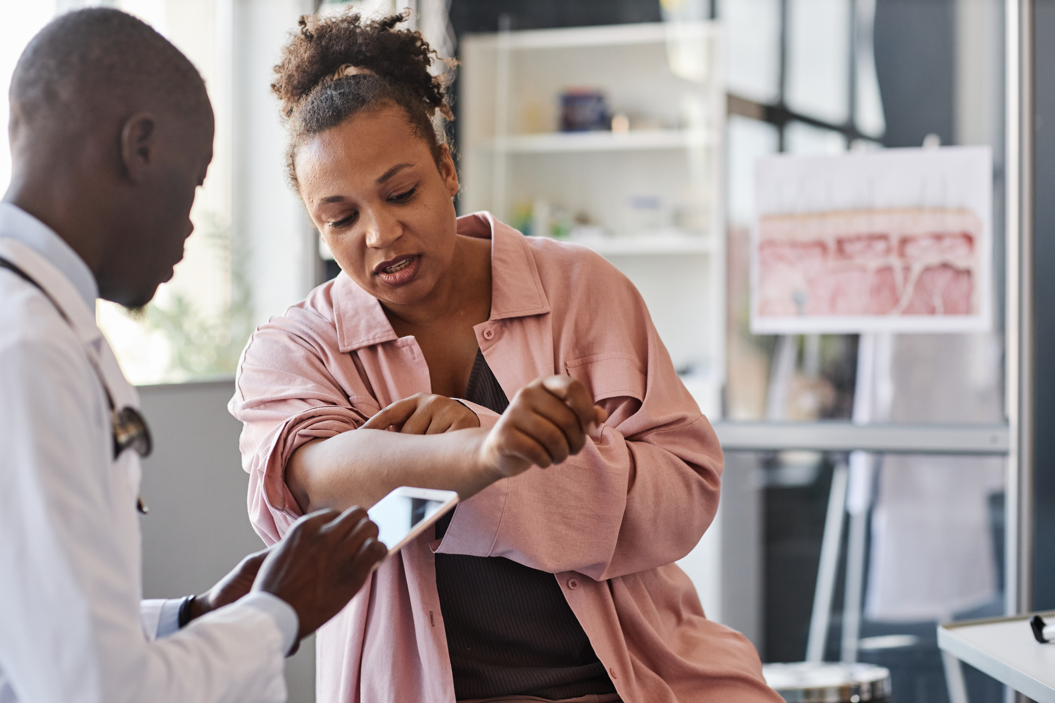 Adult African American woman suffering from psoriasis visiting dermatologist in clinic and pointing on rash spots on arms copy space