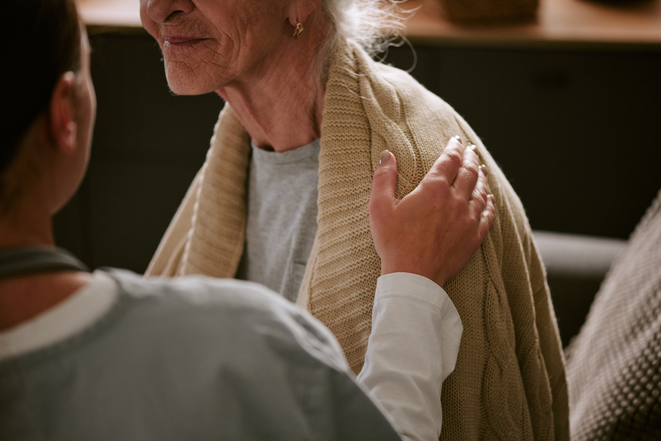 Senior Caucasian woman sitting while young adult Caucasian female caregiver gently placing hand on her shoulder, showing support and care in healthcare or home setting