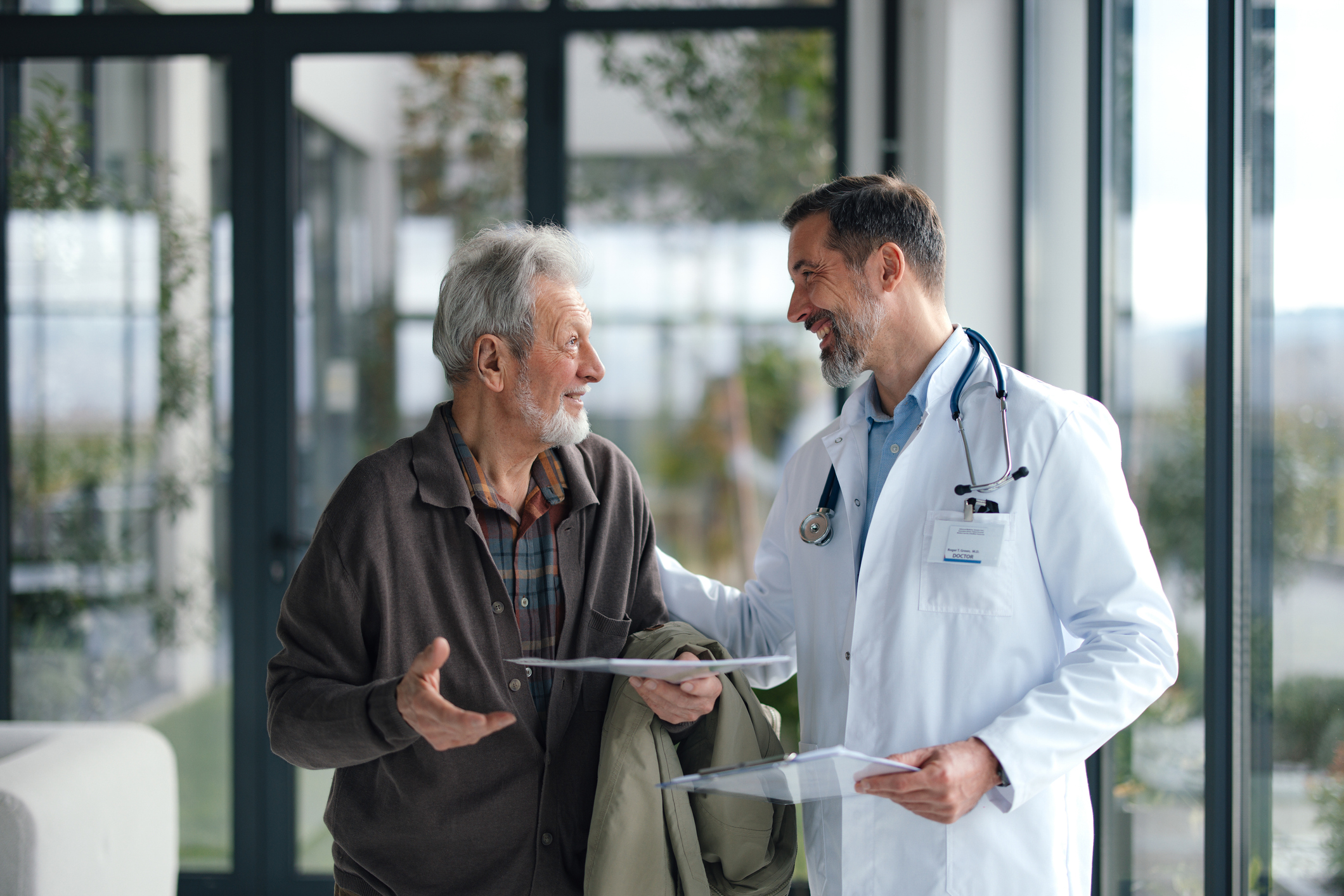 A warm interaction between an elderly patient and a doctor in a modern hospital lobby. They smile while exchanging papers, conveying trust, care, and collaborative health discussion.