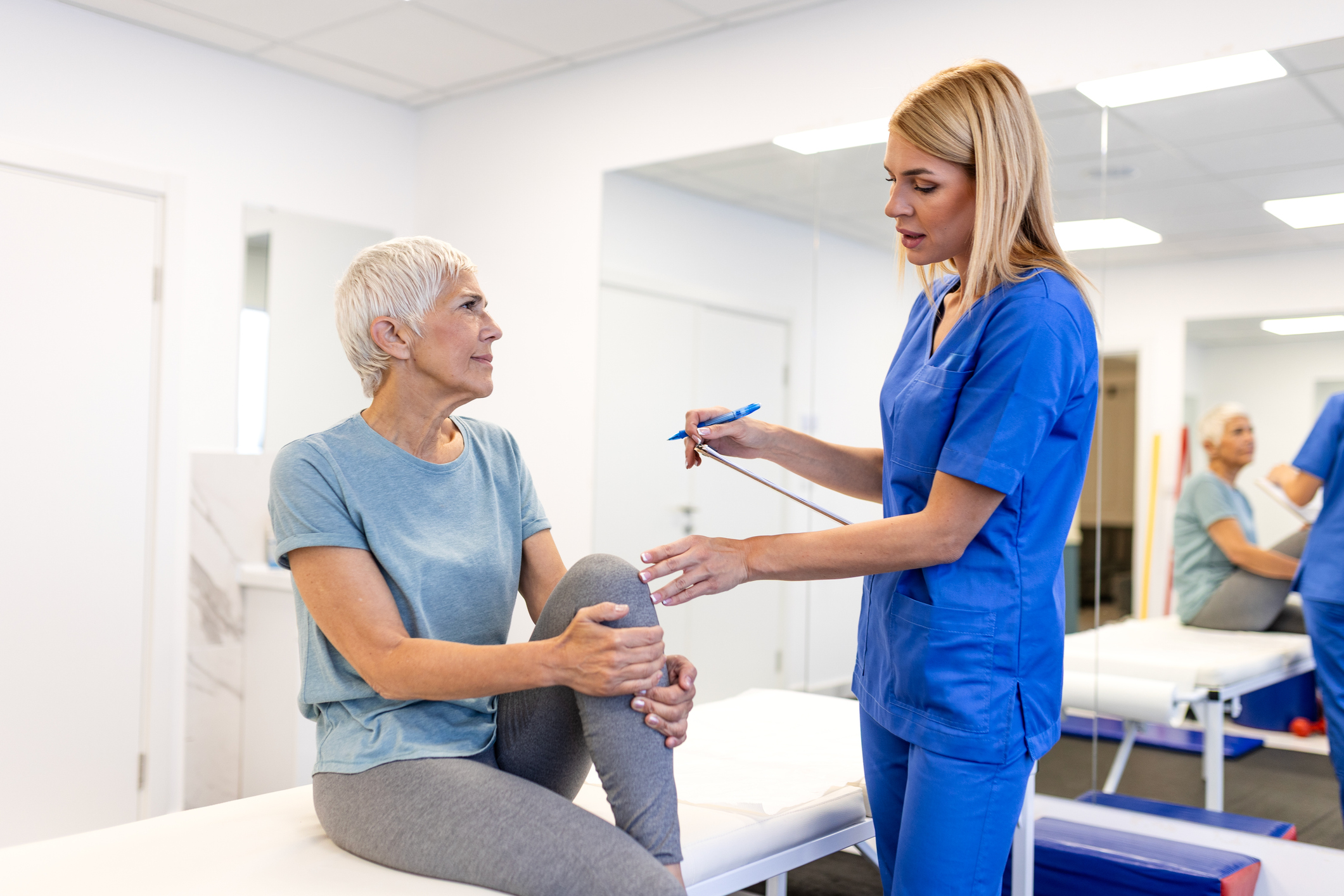 Senior Woman Discussing Knee Pain with Physiotherapist in Clinic. Healthcare and Medicine Concept. Female Patient Sitting on Examination Bed while Doctor Takes Notes during Medical Consultation.